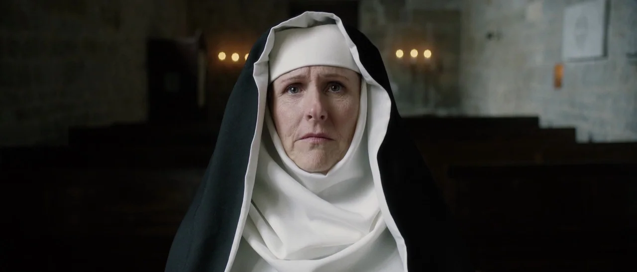 The Little Hours (2017) movie still: closeup — A stone-walled interior with wooden pews visible in the dark, out-of-focus background. A nun in…