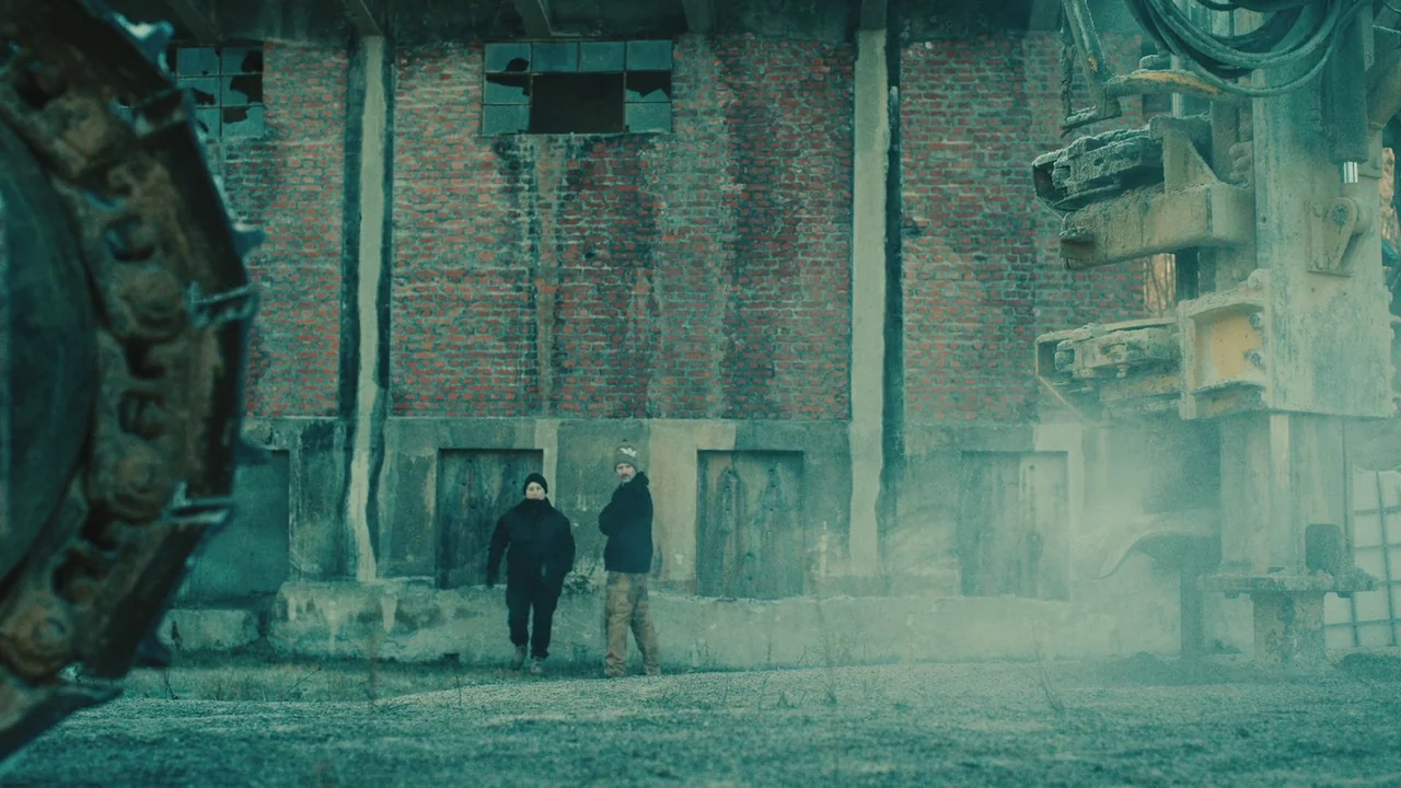 Helll Hole (2024) movie still: wide — A weathered red brick wall with broken windows stands behind two people. The figures stand in an industrial…
