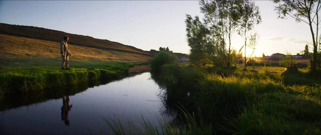 About Dry Grasses (2023) movie still: wide — A man stands near a narrow, calm river at sunset near a grassy bank. A low hill rises in the background…