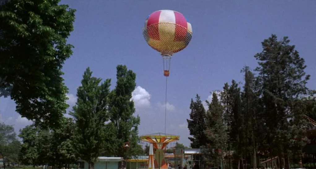 Dr. Goldfoot and the Girl Bombs (1966) movie still: wide — An amusement park with tall green trees and rides under a clear blue sky. A colorful hot…
