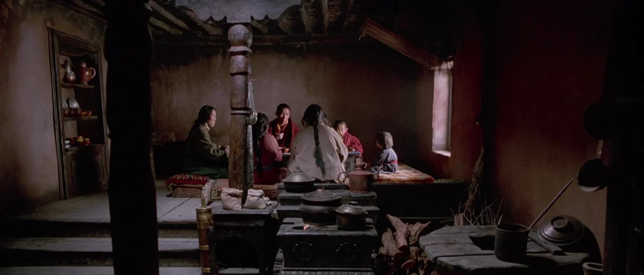 Kundun (1997) movie still: wide — A dimly lit room with mud walls contains a stone stove and wooden beams. A group sits on floor mats eating…
