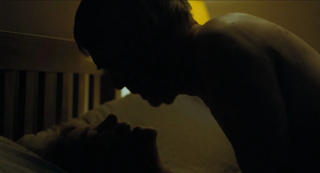 45 Years (2015) movie still: medium — A dimly lit room contains a bed with a wooden headboard. One shirtless person leans over another person lying…