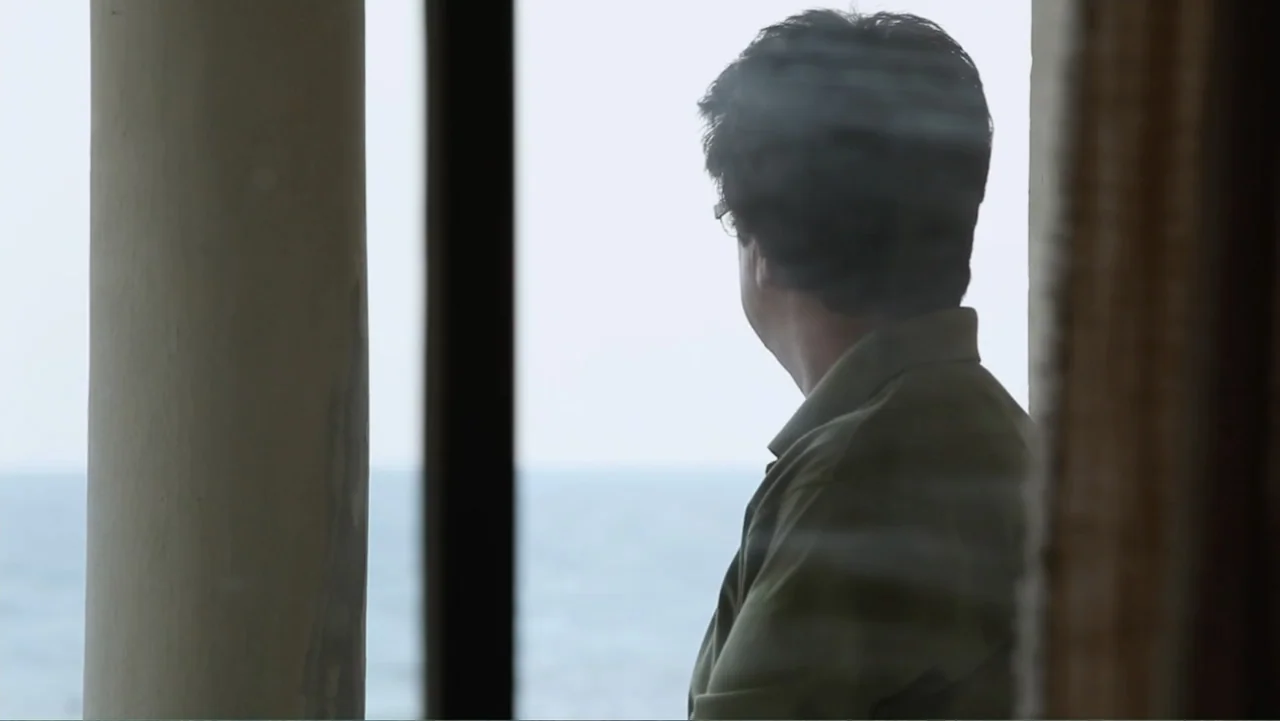 Closed Curtain (2013) movie still: medium — A man stands inside a room peering through a window towards the distant sea. A thick vertical pillar…