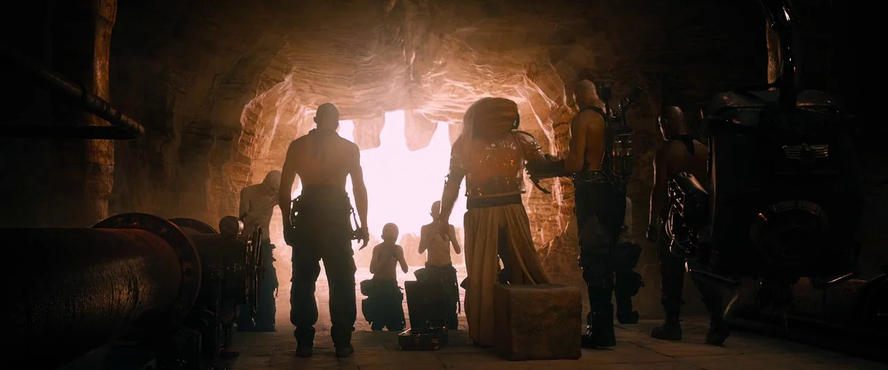 Mad Max: Fury Road (2015) movie still: wide — A dark rocky cavern opens to an intensely bright light at the rear. Several muscular figures stand in…