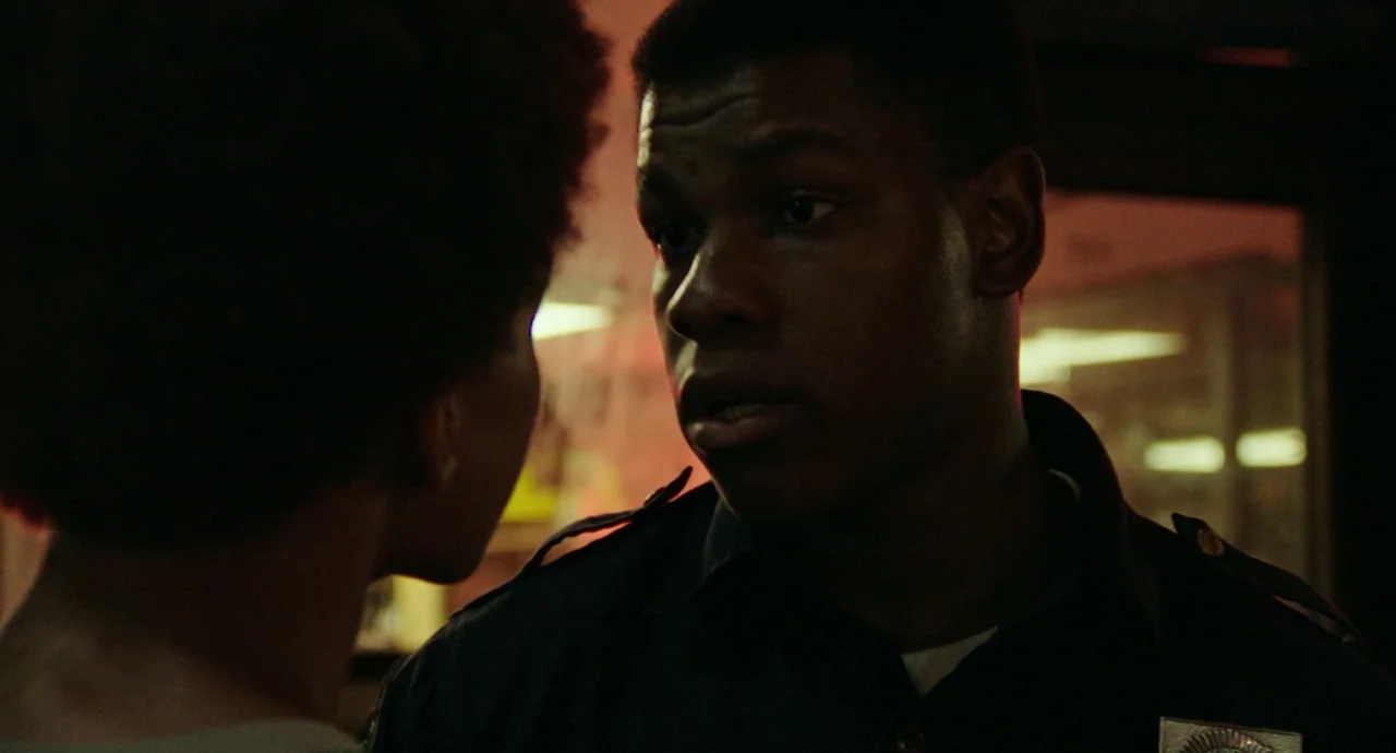 Detroit (2017) movie still: closeup — A dark, dimly lit setting with warm neon lighting reflecting on surfaces. A man wearing a police uniform faces…