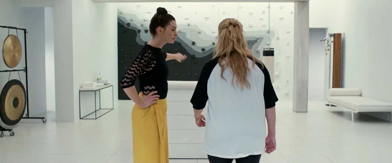 The Hustle (2019) movie still: fullbody — A stark white fitness studio contains a climbing wall and metallic gongs in corners. One woman in a black…