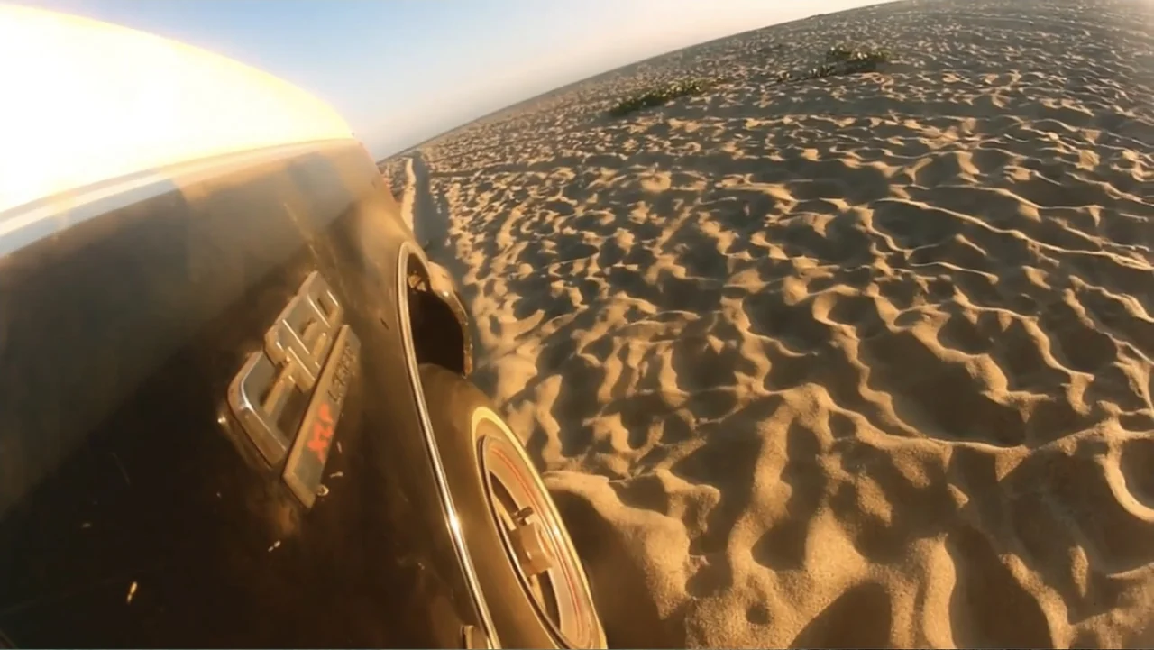 El sueño (2020) movie still: wide — A vast sandy beach at golden hour stretches toward the distant horizon line. A black vehicle tire moves forward…