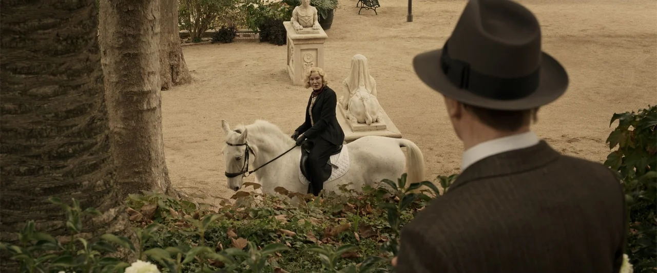 Marlowe (2023) movie still: medium — A sandy courtyard features stone statues and sparse trees under bright daylight. A woman rides a white horse…