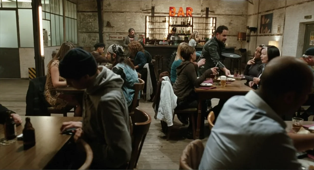 Soul Kitchen (2009) movie still: wide — A spacious bar with brick walls and rustic wooden furniture is filled with patrons. Multiple small groups…