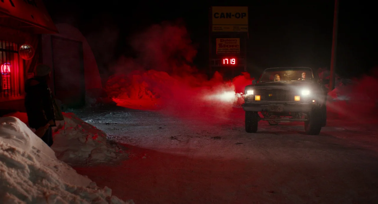 My Animal (2023) movie still: wide — A dark, snow-covered parking lot at night features a glowing red gas station price sign. A pickup truck with…