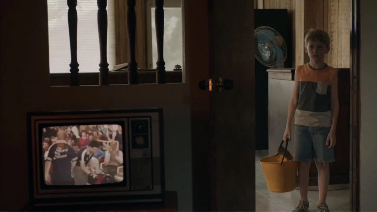 Boy (2010) movie still: medium — A domestic interior with wood paneling and a vintage television displaying a broadcast image. A young child in…