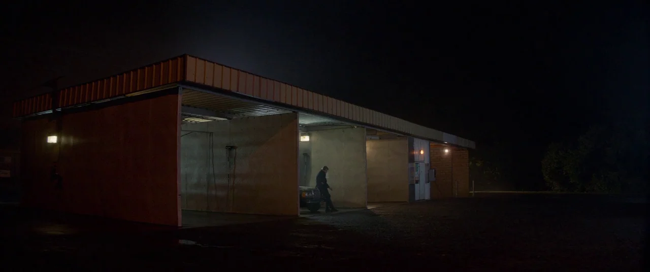 Woodshock (2017) movie still: wide — A roadside self-service car wash at night consists of concrete bays and metal roofing. A man stands alone in…
