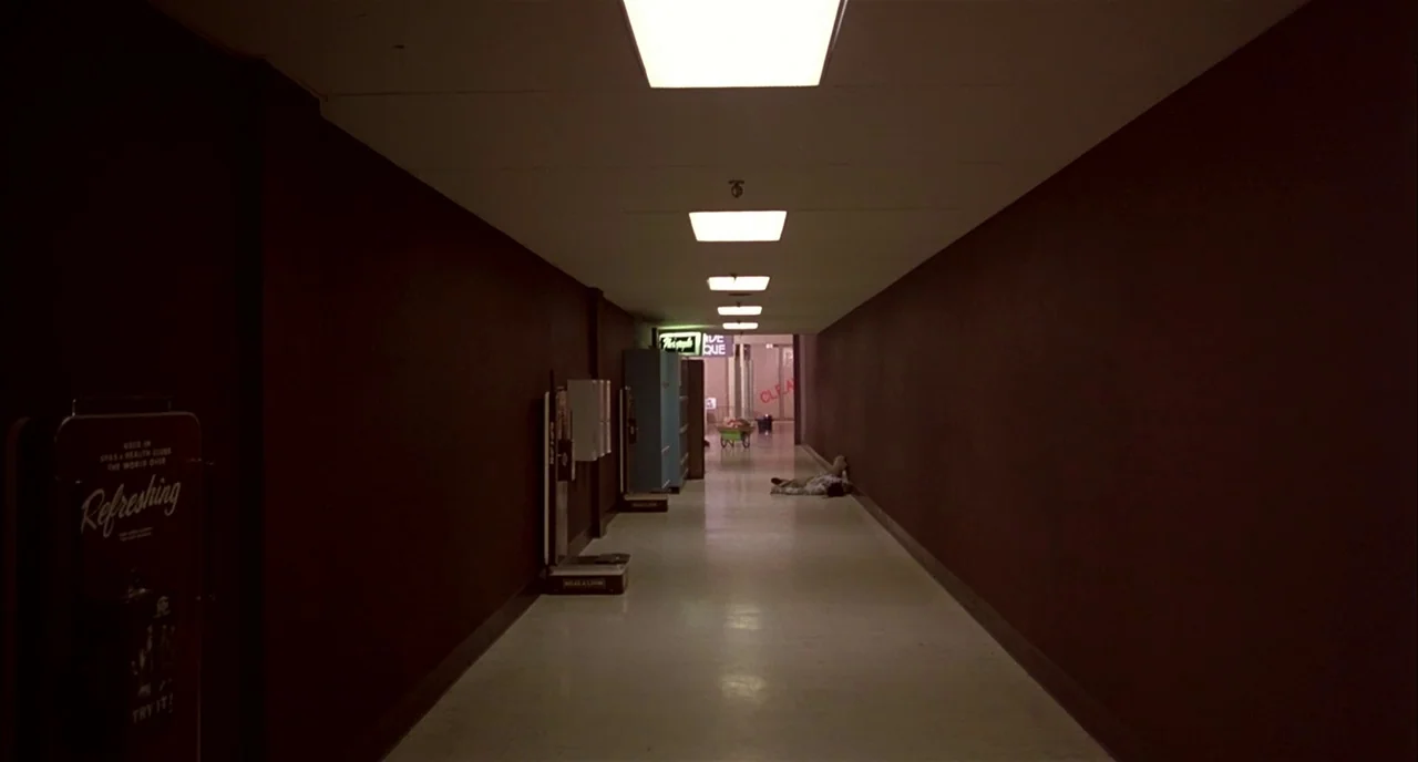 Dawn of the Dead (1978) movie still: wide — A long, dark mall corridor features deep brown walls and a sterile white floor. A single person lies…