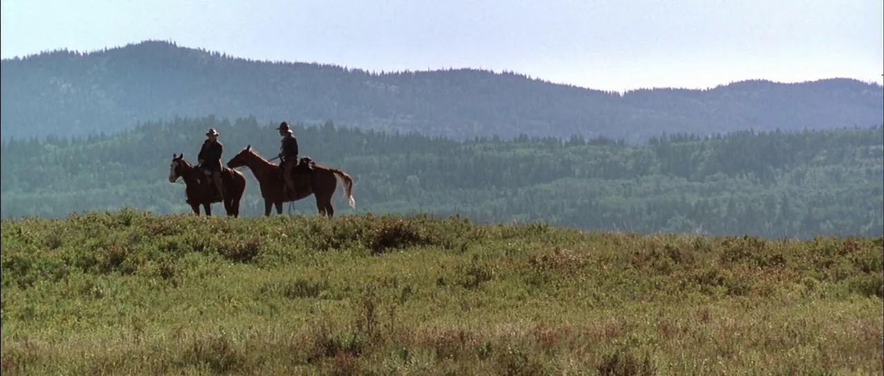 Open Range (2003) movie still: wide — A grassy expanse leads up to a forested ridge under a clear pale sky. Two riders on horseback pause on the…