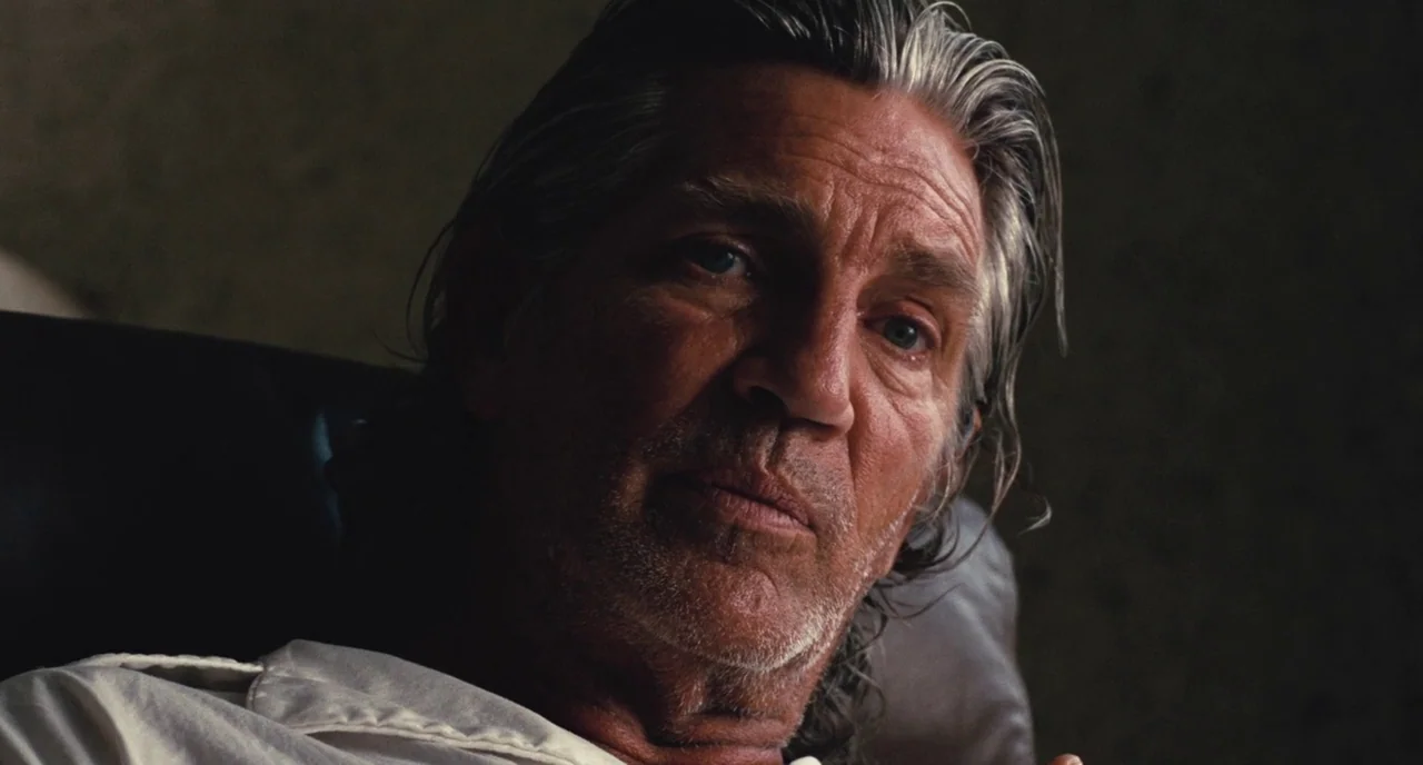 Inherent Vice (2014) movie still: closeup — A dimly lit interior space with a plain textured wall as the backdrop. An older man with grey hair sits…