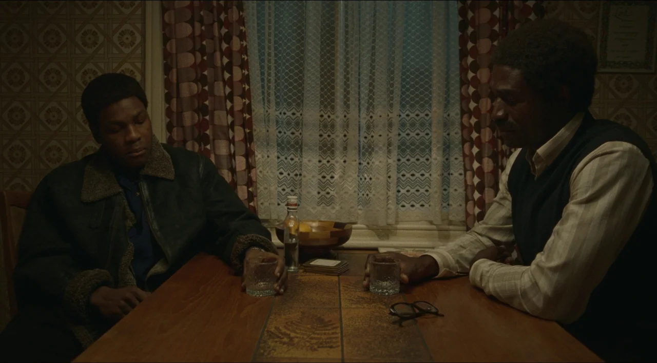 Red, White & Blue (2020) movie still: medium — A 1970s dining area with patterned wallpaper and lace-curtained windows at night. Two men sit across…