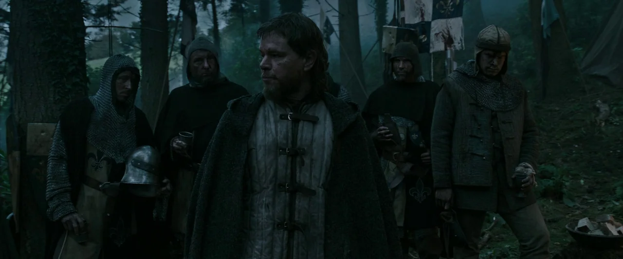 The Last Duel (2021) movie still: medium — A group of medieval soldiers gather in a dark, overgrown forest clearing. They wear padded armor,…