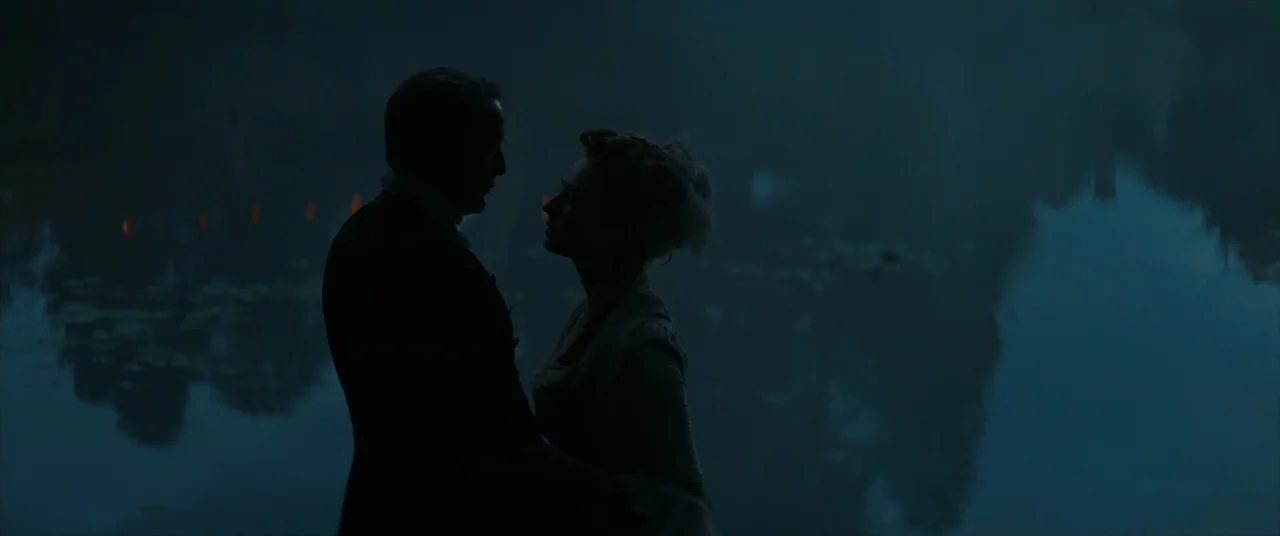 The Promised Land (2023) movie still: medium — A man and a woman stand close to each other beside a dark lake. They are silhouetted against the dim,…