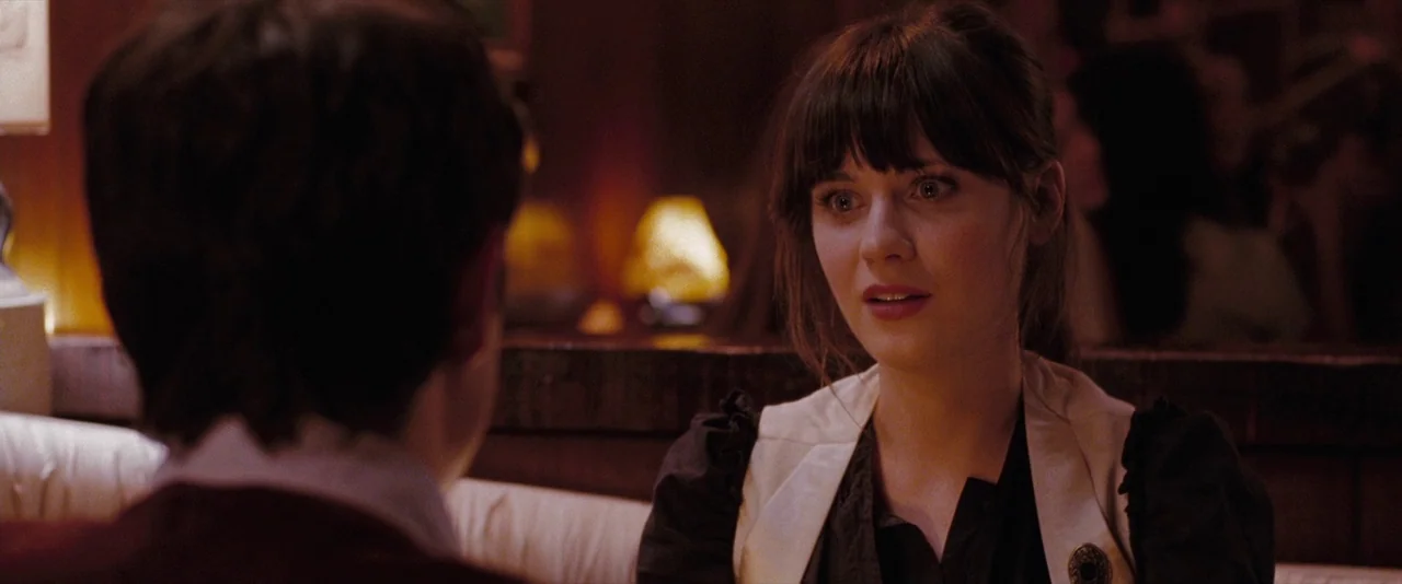 500 Days of Summer (2009) movie still: medium — A dark, wood-paneled booth setting is lit by distant glowing table lamps. A young woman with dark…