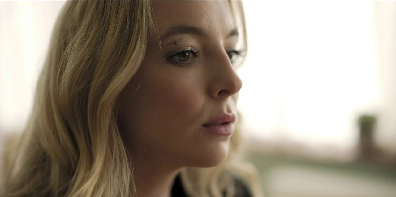 Help (2021) movie still: closeup — A close-up view of a young woman with blonde hair against a bright background. She turns her head slightly while…