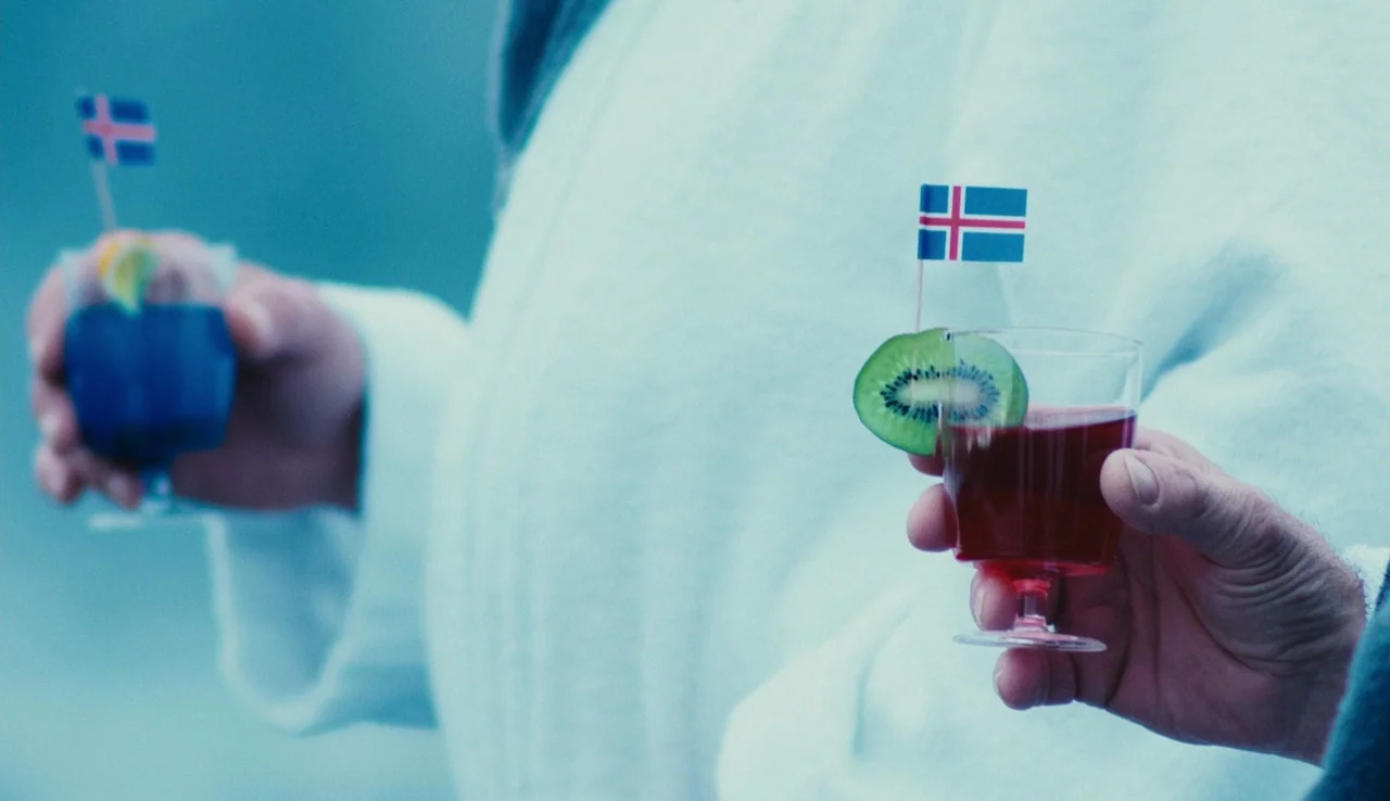 Land Ho! (2014) movie still: medium — A close-up view shows two hands holding small glasses decorated with miniature Icelandic flags. One glass…