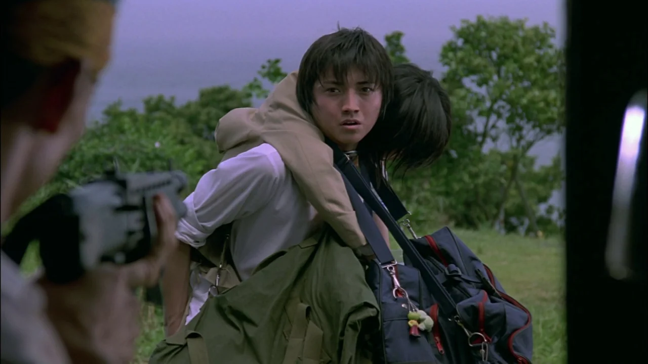 Battle Royale (2000) movie still: medium — An open grassy field under an overcast sky borders a body of water. A young man carries an unconscious…