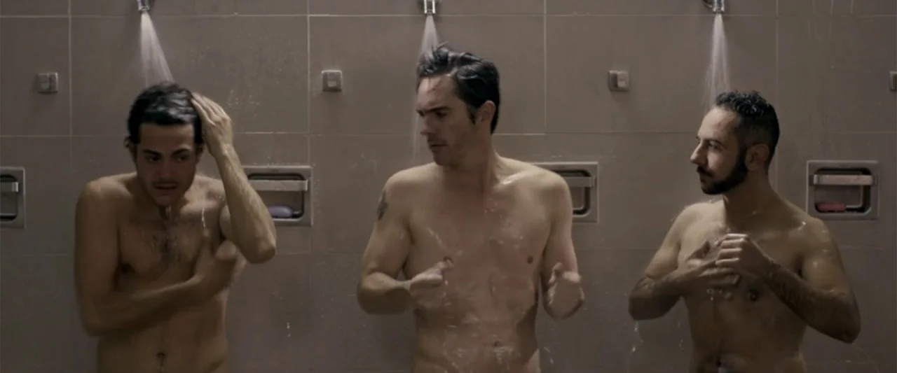 Do It Like An Hombre (2017) movie still: medium — Three shirtless men stand side-by-side in a tiled public shower facility. Each man operates a…