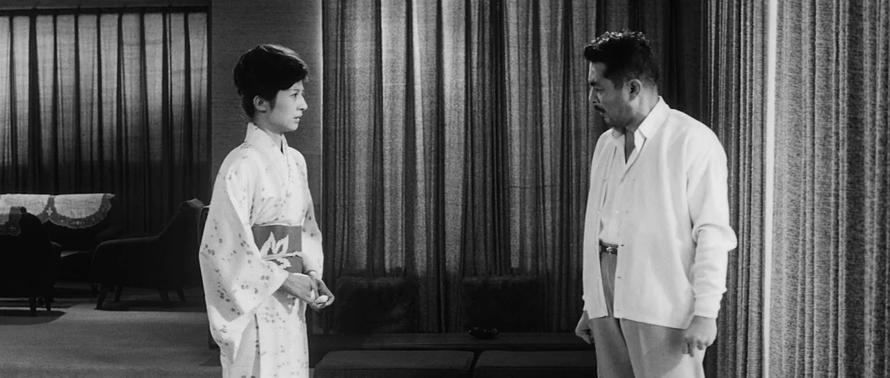 High and Low (1963) movie still: medium — A mid-century modern room features floor-to-ceiling pleated curtains and dark carpeted floors. A woman in…