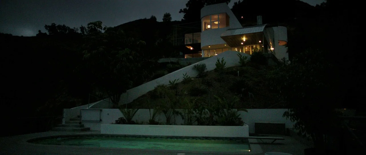 The Canyons (2013) movie still: establishing — A modern hillside residence with white walls sits above a pool at night. Interior lights shine…