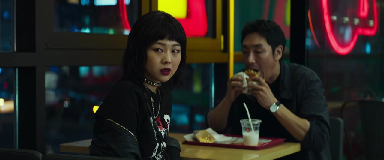 Believer (2018) movie still: medium — A dark, modern restaurant interior at night features bright red and yellow neon lights. A young woman and a…