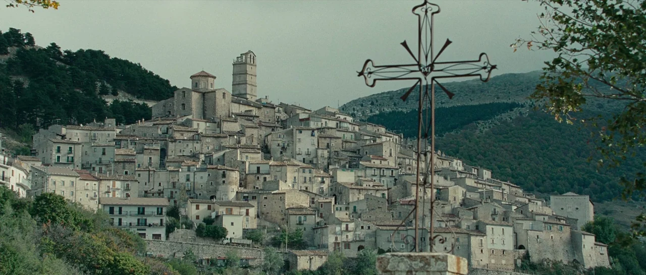 The American (2010) movie still: establishing — A dense hillside village features tiered stone houses climbing up a steep mountainside. A prominent…