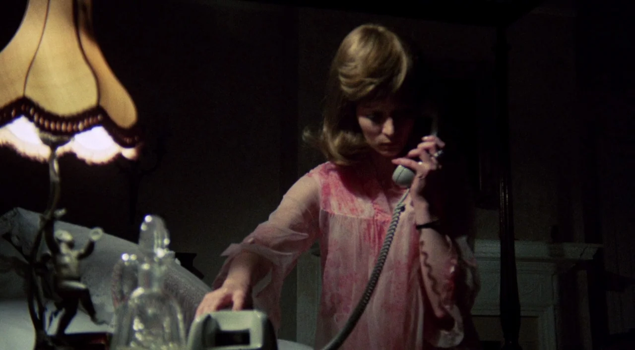 The Iguana with the Tongue of Fire (1971) movie still: medium — A woman stands in a dim room near a vintage lamp. She holds a telephone receiver…