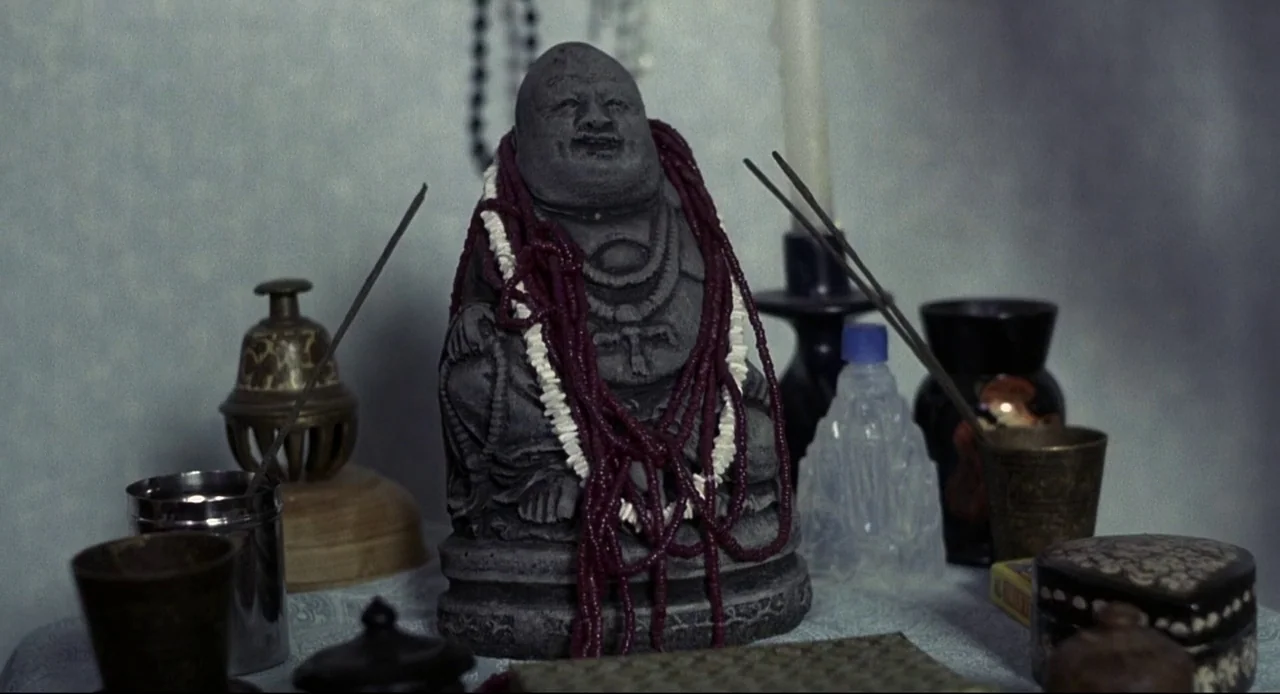 Naked (1993) movie still: closeup — A stone statue sits on a table covered in various ritualistic items and beads. The scene is static with no…