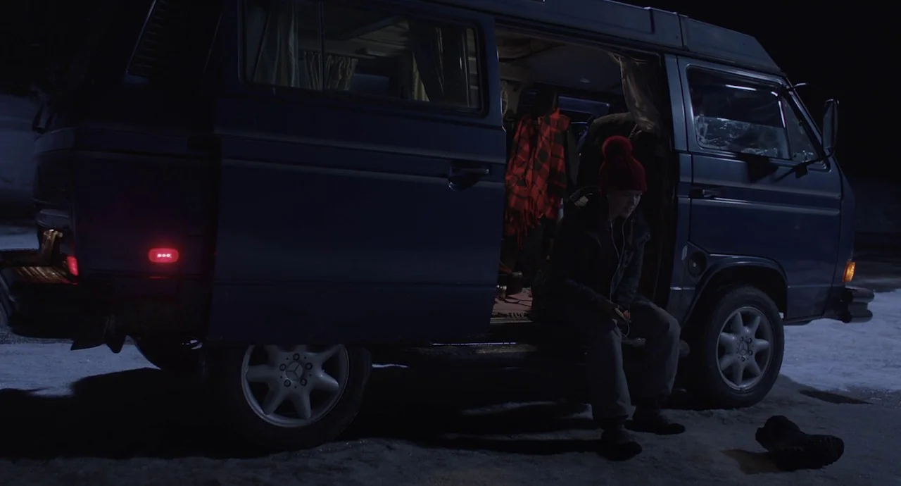 The Wolf of Snow Hollow (2020) movie still: fullbody — A dark blue van with an open sliding door sits on snow at night. A young woman in a red…