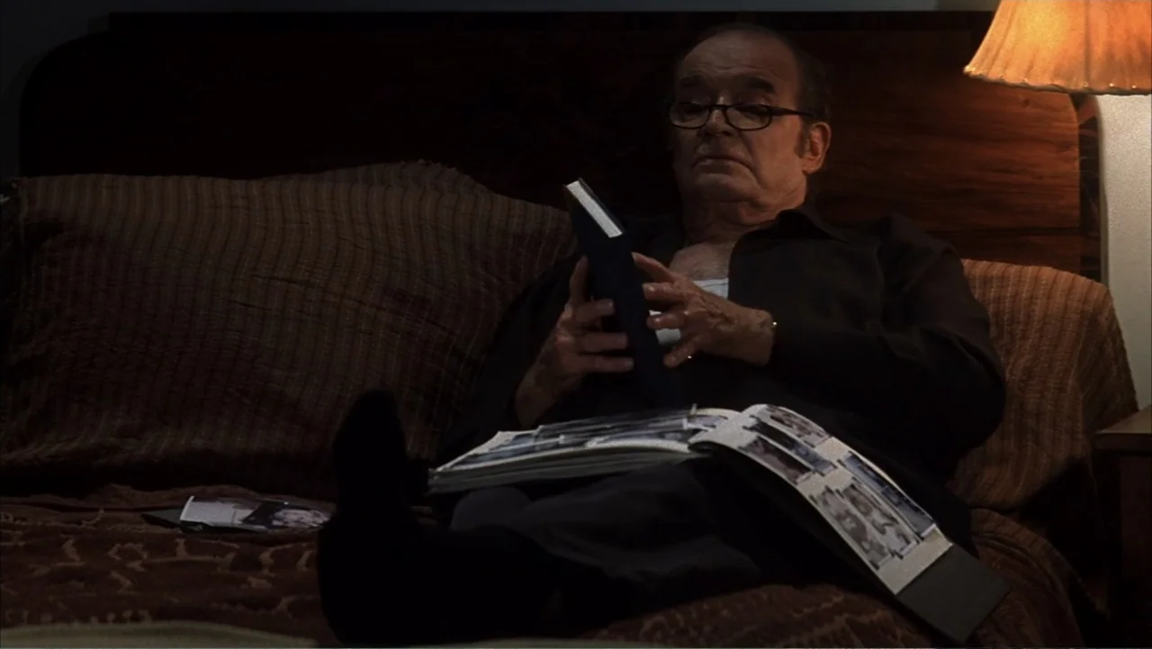 The Notebook (2004) movie still: medium — An elderly man sits in a dimly lit bedroom with a large wooden headboard. He is focused on a book and…
