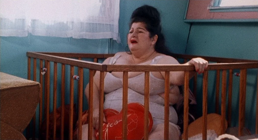 Pink Flamingos (1972) movie still: medium — A woman sits inside a wooden infant crib against a turquoise-painted wall. She holds a red heart-shaped…