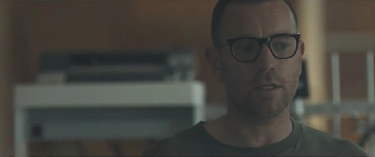 Zoe (2018) movie still: closeup — A man stands inside a softly lit office with neutral-toned walls. He is wearing glasses and a plain olive green…