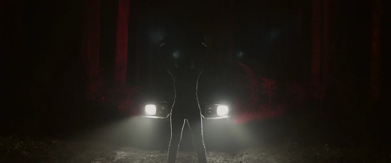 Impetigore (2019) movie still: fullbody — A dirt path through a dark woodland at night is illuminated by car headlights. A person stands centrally…