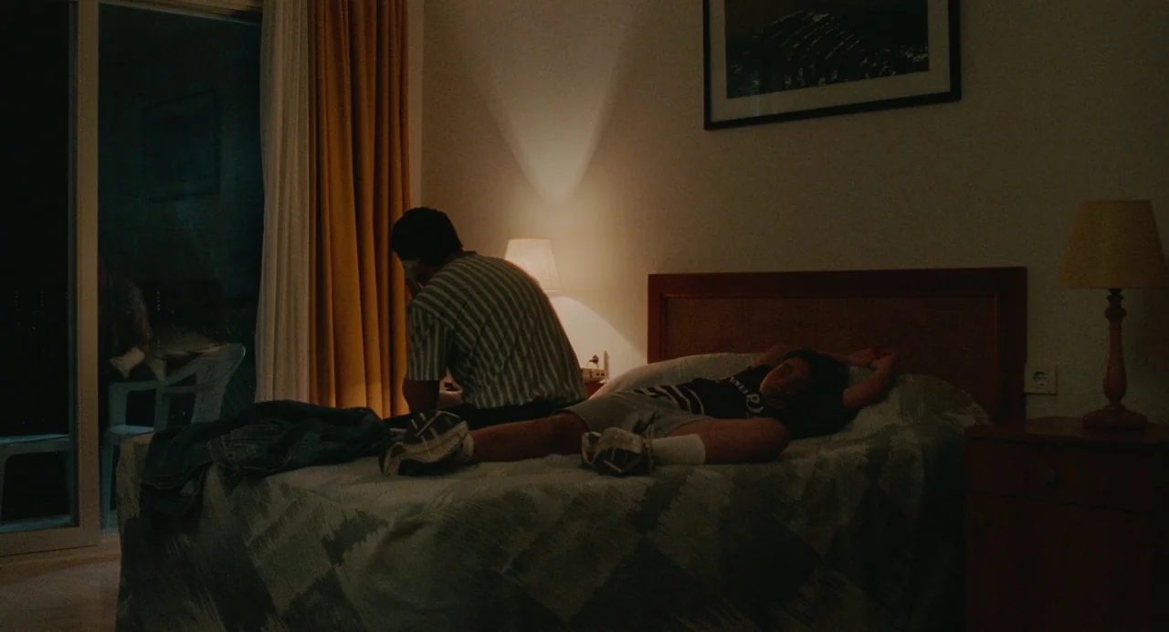 Aftersun (2022) movie still: medium — A dimly lit hotel room features a bed with patterned covers and a balcony door. A man sits facing away from…