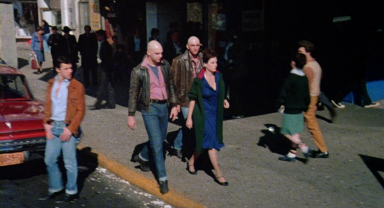 The Wanderers (1979) movie still: fullbody — A sunny 1970s urban sidewalk with a red vintage car parked on the left. Three people walk in a line…