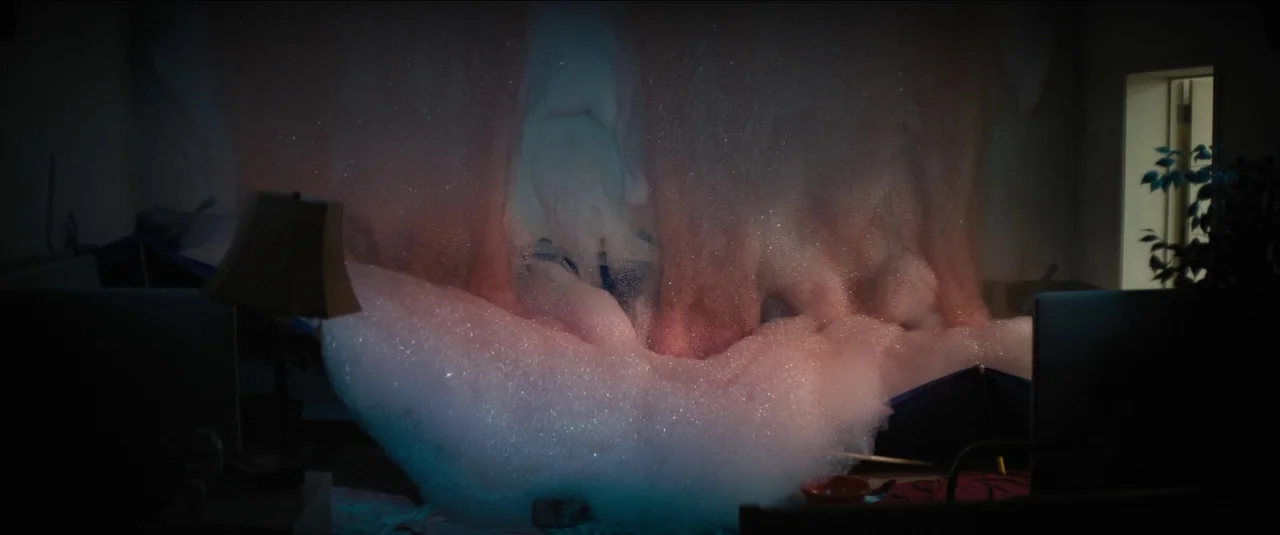 Kajillionaire (2020) movie still: wide — A dark, dimly lit room is rapidly filling with a massive mound of foam. The thick white and pinkish bubbles…