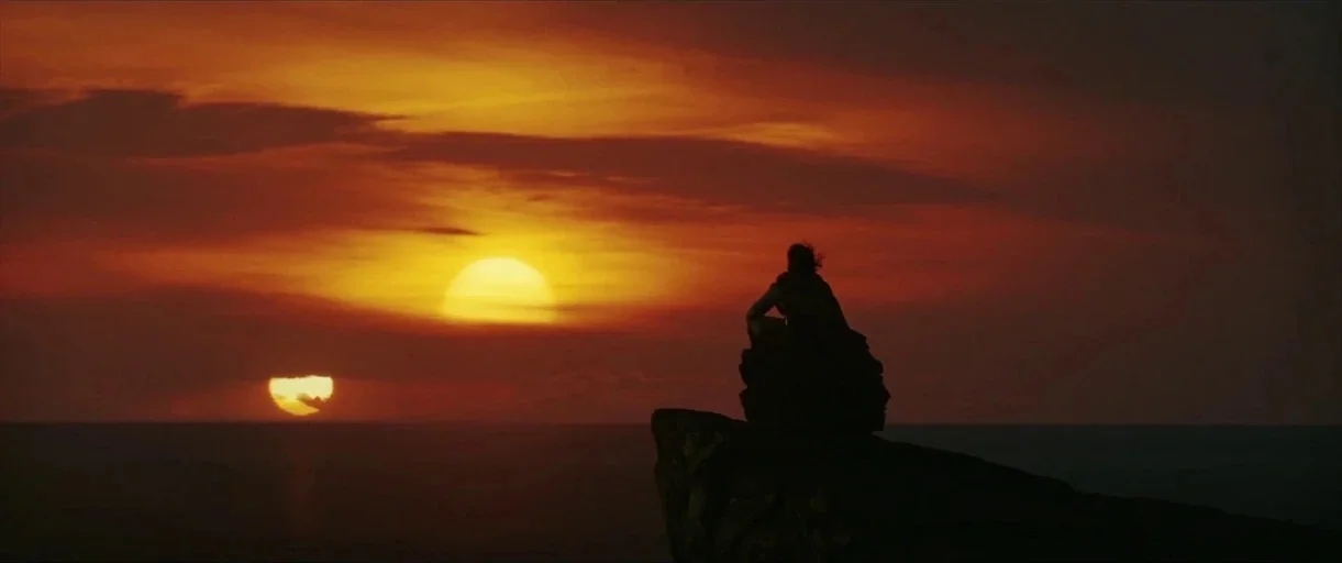Star Wars: The Last Jedi (2017) movie still: wide — A rugged cliff edge overlooks an expansive desert landscape under a double sunset. A lone person…