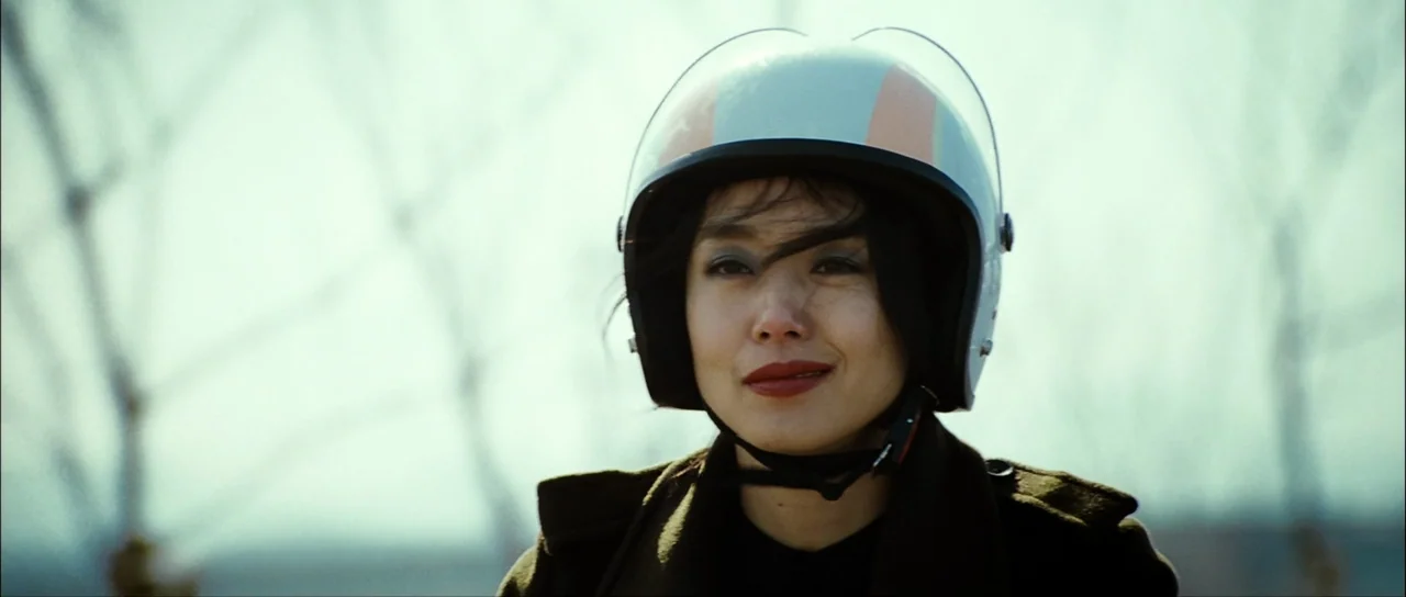 The Housemaid (2010) (2010) movie still: closeup — A woman wearing a white motorcycle helmet looks towards the camera outdoors. Bare tree branches…