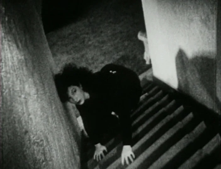 Meshes of the Afternoon (1943) movie still: fullbody — A dark, narrow indoor staircase with a rough wall on one side. A woman crawls upward on her…