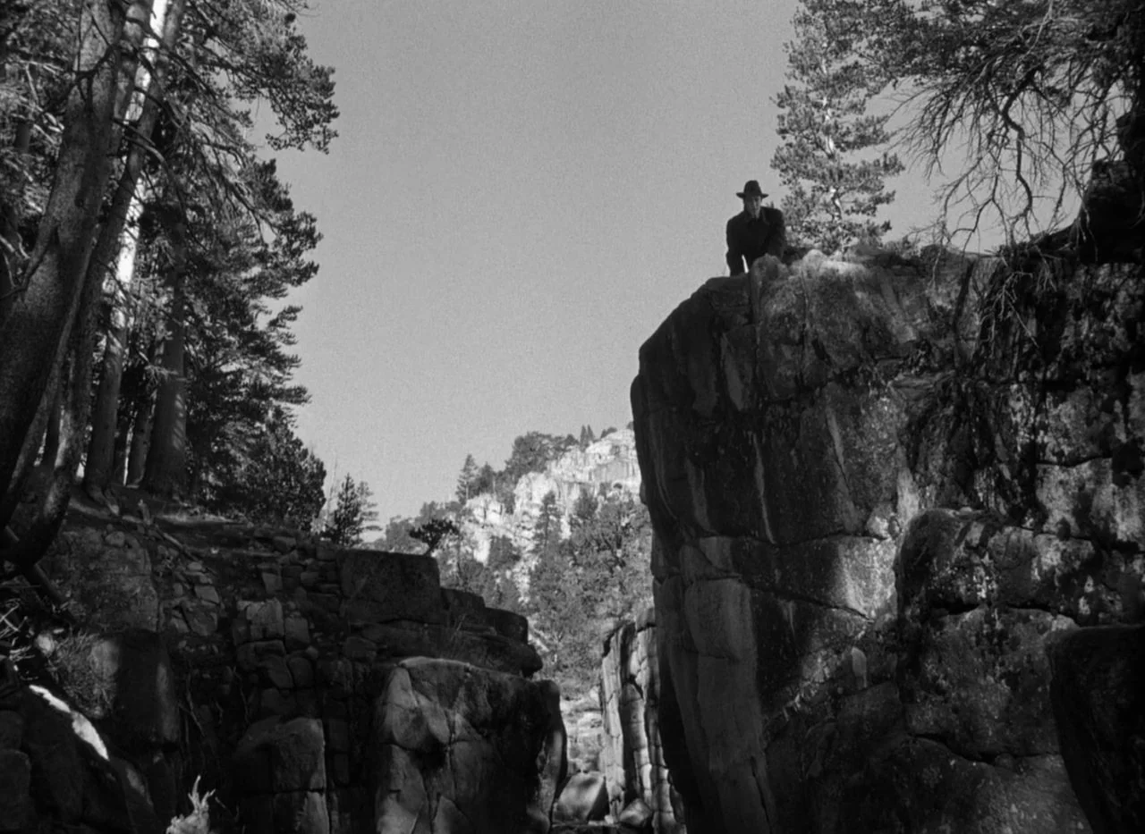 Out of the Past (1947) movie still: wide — A deep rocky canyon is flanked by towering stone walls and evergreen trees. A lone man wearing a brimmed…