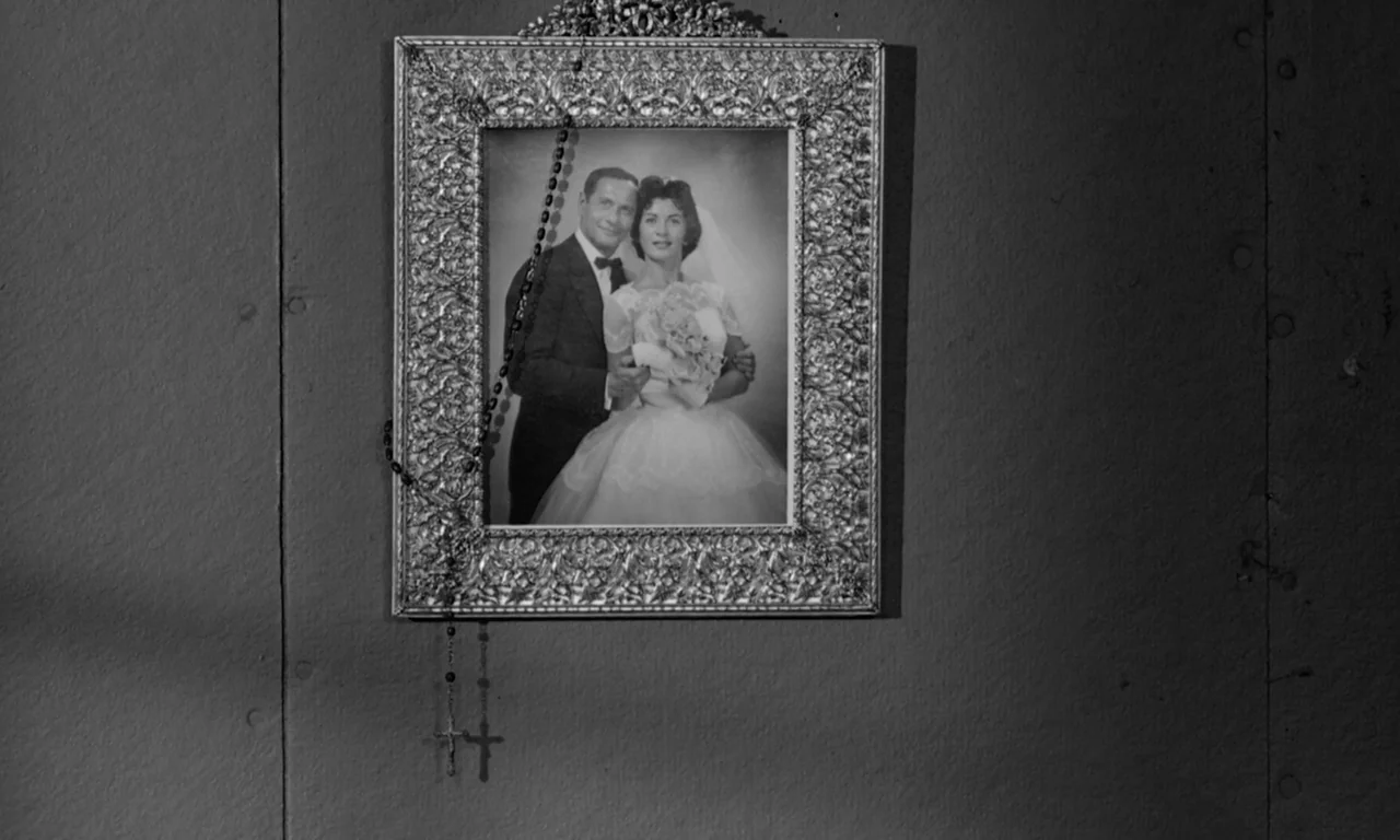 The Misfits (1961) movie still: closeup — A framed 1950s wedding photograph is mounted on a plain, dark-colored wall. A black rosary is draped over…