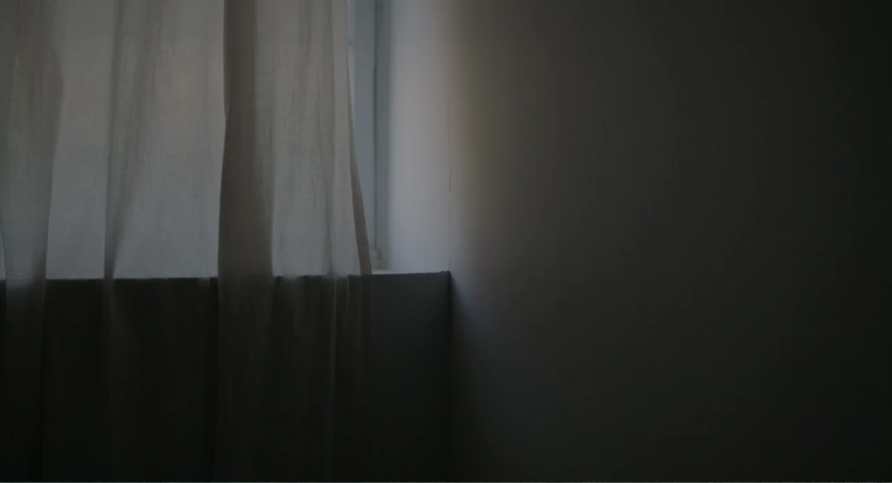 This Closeness (2024) movie still: establishing — A corner of a room features a white wall and a thin, hanging curtain. Natural light from a hidden…