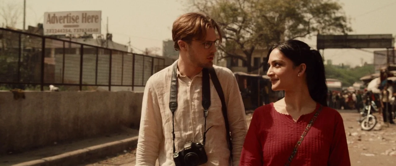 I Origins (2014) movie still: medium — A sunny city street featuring a concrete wall and an advertisement billboard behind. A man with a camera and…