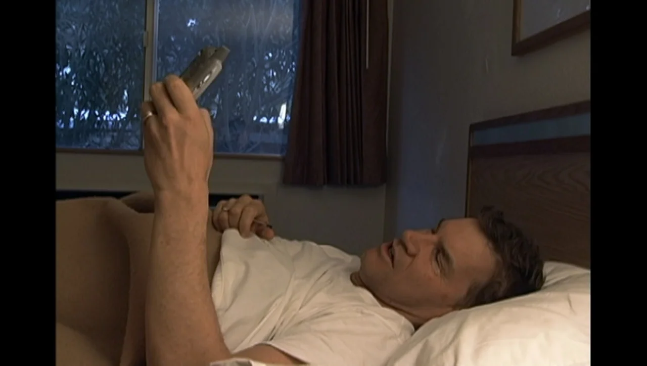 The Trials of Ted Haggard (2009) movie still: medium — A man lies in a bed with a wooden headboard in a simple motel room. The man holds a small…