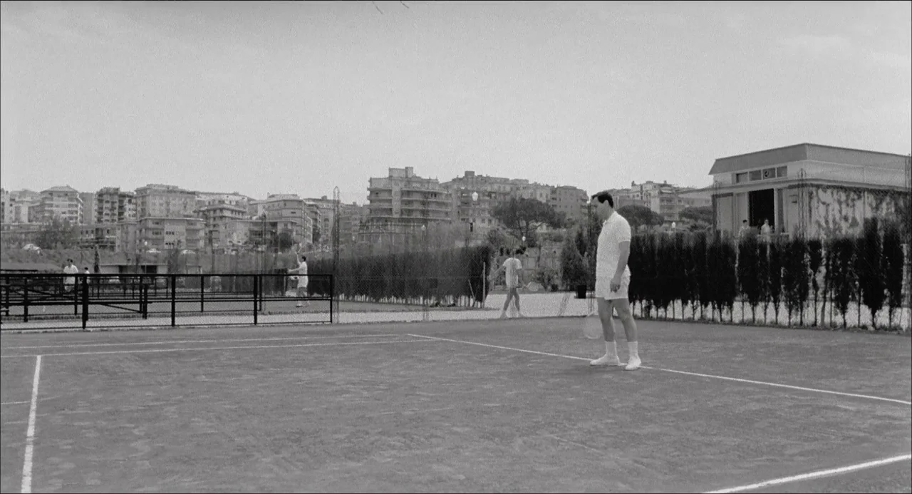 Il Boom (1963) movie still: wide — A clay tennis court sits before a background of urban apartment buildings. A man stands in the foreground on the…
